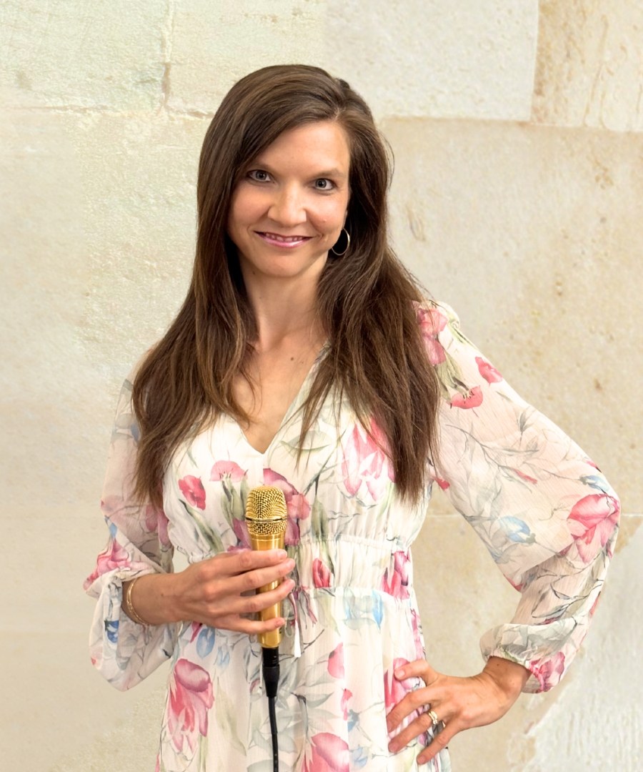 Luisa Loreen im geblümten Kleid mit goldenem Mikrofon vor einer hellen Sandsteinwand