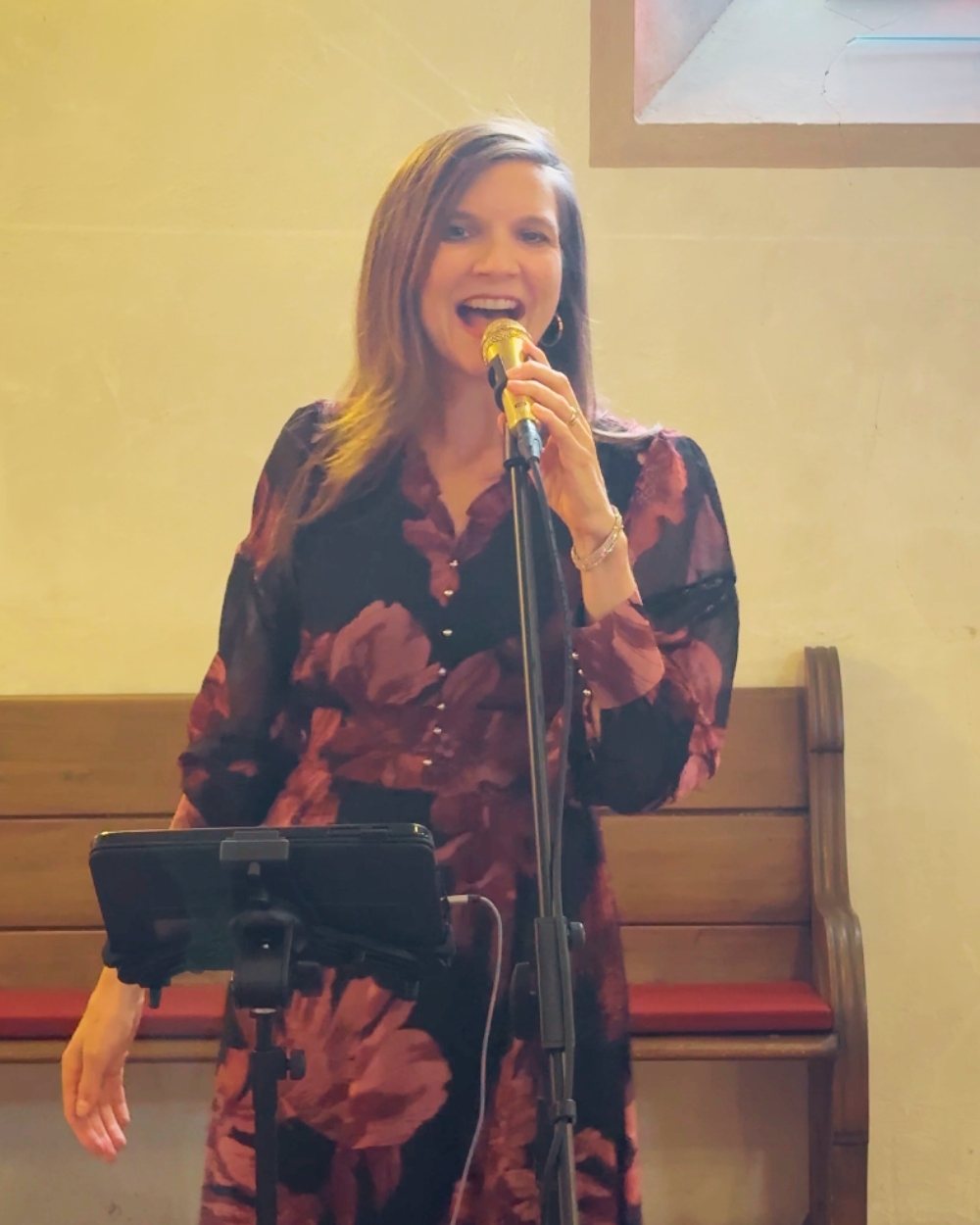 Luisa Loreen singt in einem dunklen Blumenkleid in ein goldenes Mikrofon am Stativ; hinter ihr eine helle Wand und eine Holzbank.