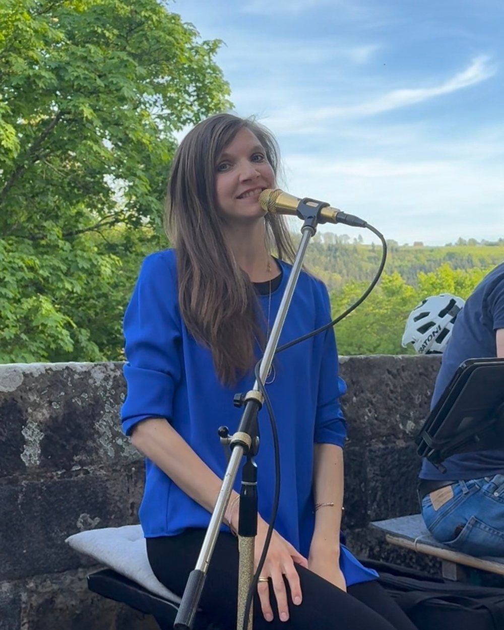 Luisa Loreen sitzt in blauer Bluse auf einer Steinmauer im Freien und singt in ein goldenes Mikrofon; dahinter Bäume und eine weite Landschaft.