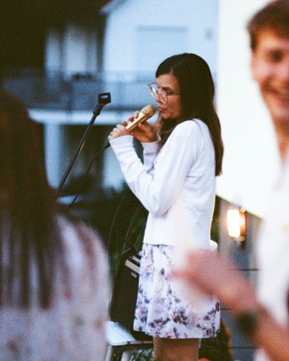 Luisa Loreen singt am Abend im Freien in ein goldenes Handmikrofon; im Vordergrund stehen unscharf einige Gäste.