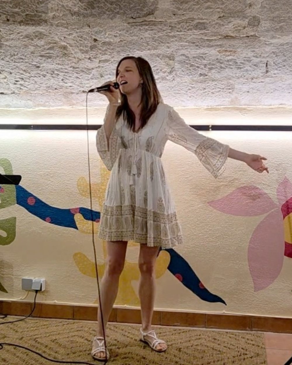 Luisa Loreen im weißen Kleid singt in ein Handmikrofon vor einer Wand mit bunter Blumenmalerei unter einer alten Natursteindecke.