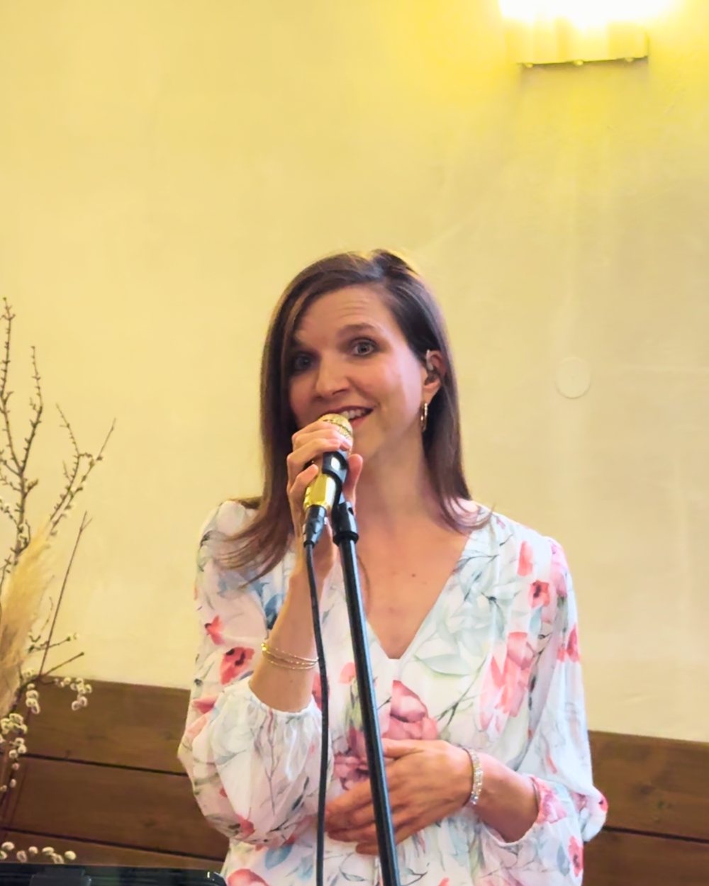 Luisa Loreen singt in einem geblümten Kleid in ein goldenes Mikrofon am Stativ, hinter ihr eine warm beleuchtete Wand mit Holzvertäfelung.