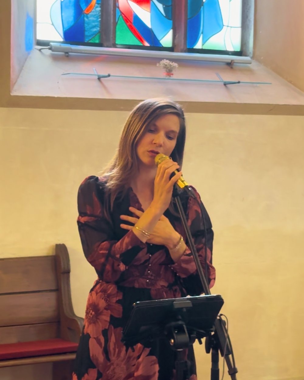 Luisa Loreen singt in einer Kirche in ein goldenes Handmikrofon, über ihr ein buntes Kirchenfenster, daneben eine Holzbank.